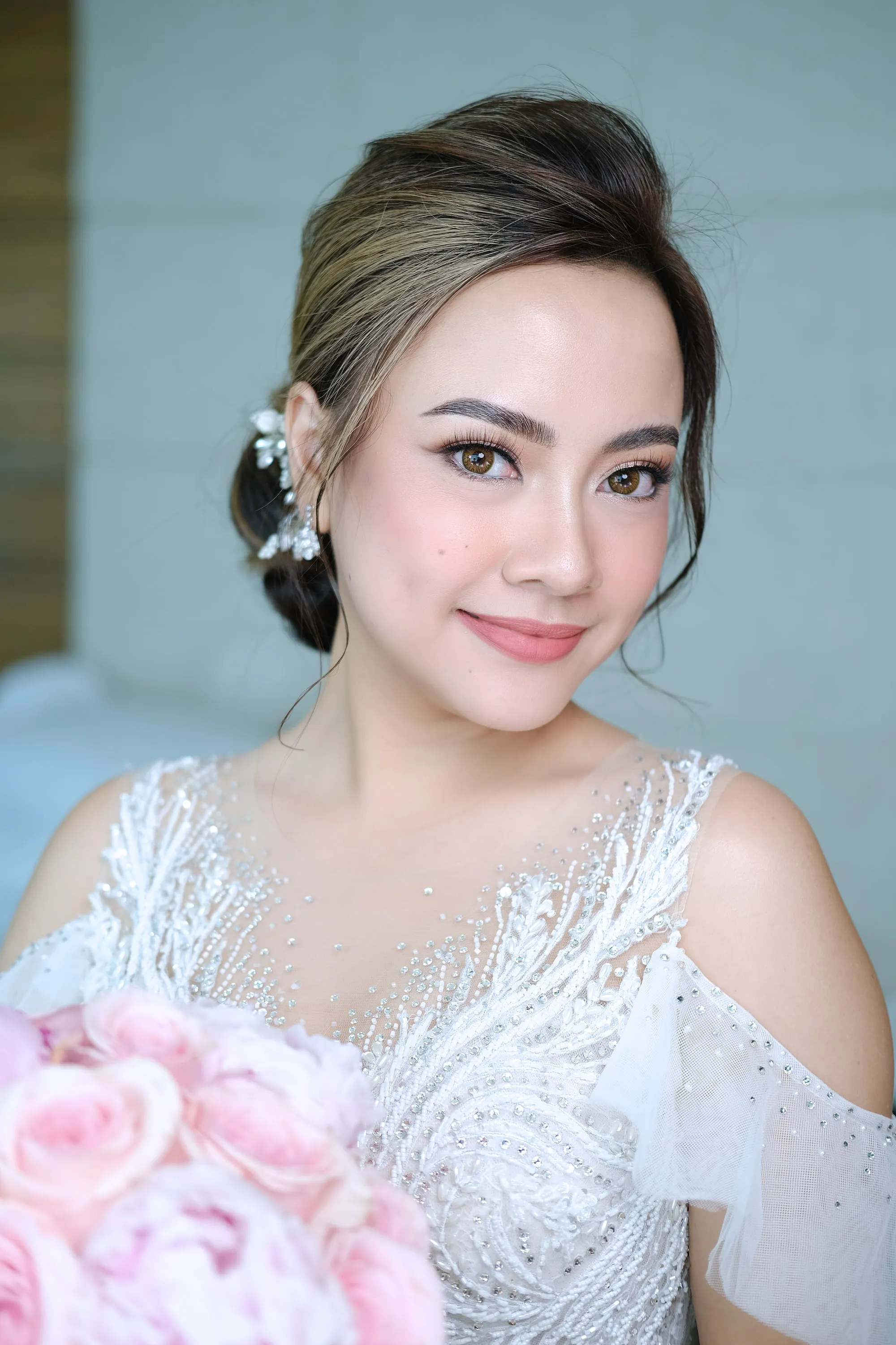 Bridal make-up with a soft rose lip and a luminous skin finish, worn with a low twisted updo, a jewelled hairpiece and a bouquet of pale pink roses.
